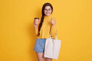 Young woman with white cotton bag and paper coffee cup in her hands looking smiling directly at camera and showing thumb up, wearing sweater and short jeans.