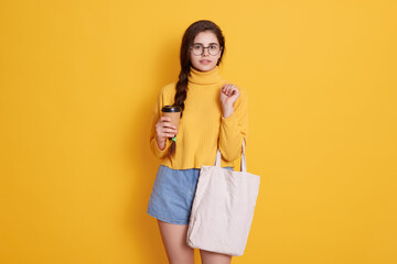 Young woman with cotton bag and paper coffee cup in her hands posing against yellow wall, wearing stylish clothing and rounded glasses, confident female going shopping.