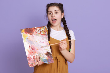 Young brunette artist woman with pigtails holding painter brush and palette over lilac background,...