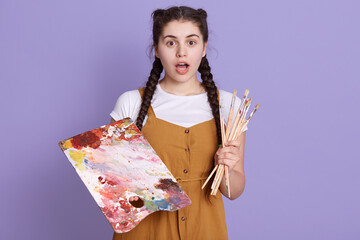 Young talented woman artist with brush and paints posing isolated over lilac background with opened...