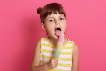 Little girl eating fruit ice cream widely opened mouth, looking at camera, wearing striped dress, has dark hair and two knots, posing isolated over pink background.