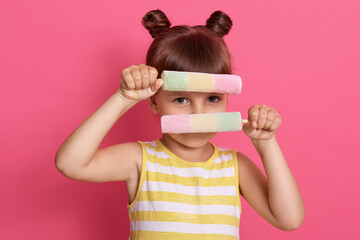 Caucasian child hiding behind two fruit ice creams, wearing summer clothes, small dark haired girl...