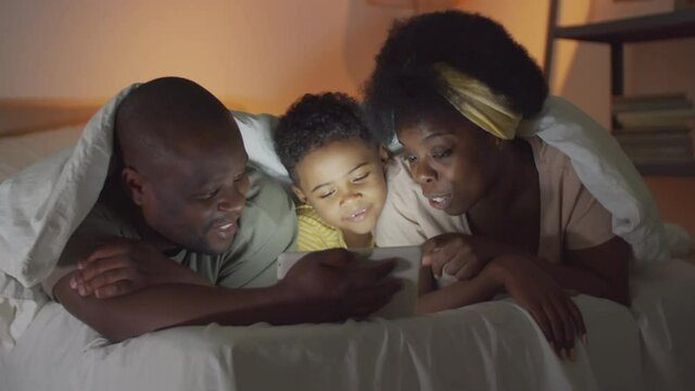 Happy African American mother, father and little son lying together under blanket on bed, watching funny cartoon on digital tablet, smiling and talking before going to bed in the evening