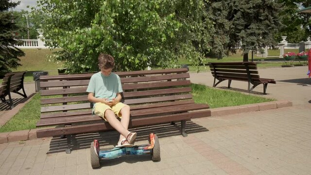 A handsome guy sits on a bench in a summer park, his feet are on a gyroscooter.