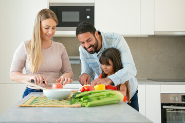 Cheerful mom and dad teaching kid to cook. Young couple and their girl cutting fresh fruits and vegetables for salad at kitchen counter. Healthy nutrition or lifestyle concept