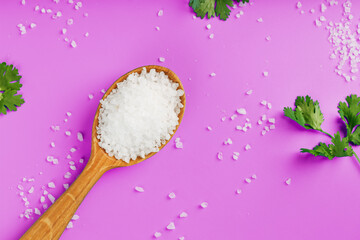 Large white sea salt in a wooden spoon on a pink background, with salt and herbs scattered around the edges.