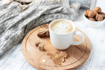 White mug with cappuccino, knitted scarf and garland on wooden table. Autumn mood. Cozy autumn composition. Hygge concept