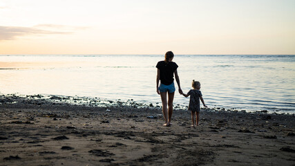 young mother and little child are walking on the sandy beach and admire the sunset