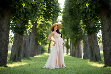 Pregnant woman posing in a beige dress on a background of green trees.