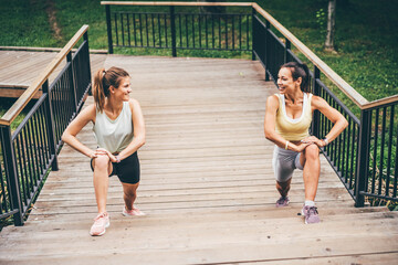 Young women with ponytails in tracksuits do stretching sports exercises on wooden staircase in green public garden on warm summer day