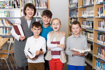 Young female librarian posing together with happy children near bookcase in school library