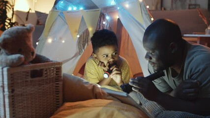 Happy African American father holding flashlight and reading fairy tale to cheerful little son while he lying in teepee tent decorated with lights in dark room at night - Powered by Adobe