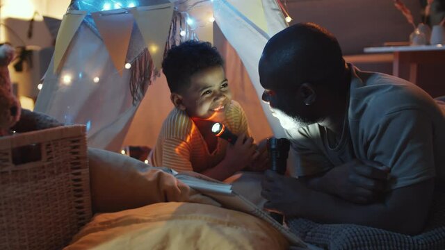 Joyous African American dad and little son holding flashlights, reading book, chatting and laughing while he lying in teepee tent decorated with lights in dark room in the evening