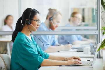 Fototapeta premium Asian young operator in uniform and headset sitting at the table and typing on laptop at hospital