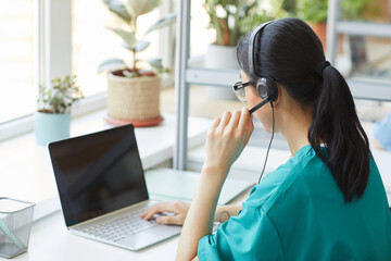 Rear view of young operator in headphones talking on the phone while sitting at her workplace in...
