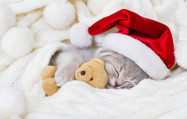 A small light tabby kitten lies on its back on a white knitted scarf in a Santa hat and holds a present with its front paws