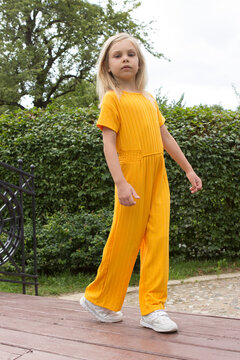 A little girl of six-seven years old with long blond hair in a plain yellow outfit stands and in a summer park on a decorative bridge and poses in full growth.