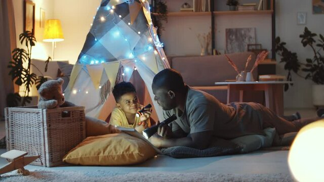 African American father holding flashlight and reading fairy tale to cute little son before going to bed while he lying on the floor under teepee tent decorated with lights in dark kids room