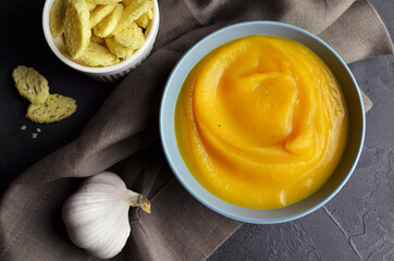 Butternut squash soup with croutons and fresh garlic in ceramic bowl on grunge black background with linen napkin. Top view