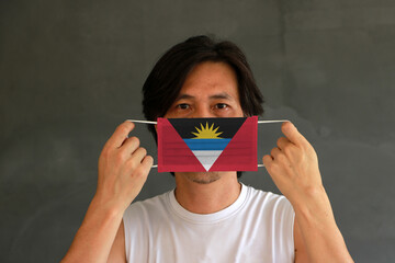 A man with Antigua and barbuda flag on hygienic mask in his hand and lifted up the front face on dark grey background. Tiny Particle or virus corona or Covid 19 protection.