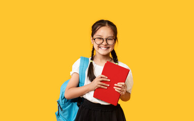 Adorable Chinese Schoolgirl With Backpack Holding Book On Yellow Background