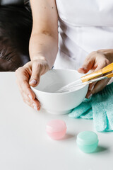Brown plastic bottle for liquid cosmetics and wooden bowl with stone sticks for face massage in woman's hands on white table. Cosmetologist holding spin care products. 