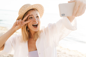 Image of happy woman showing peace sign and taking selfie on cellphone