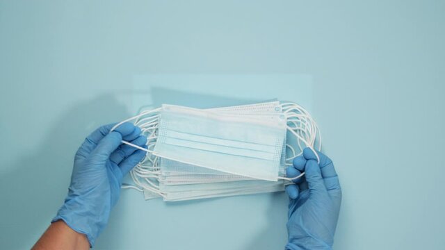 two hands in blue latex gloves lay on a blue background a stack of disposable medical face masks, protection against viruses and bacteria, top view