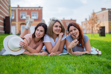 Three beautiful Caucasian young women lie on the lawn in the countryside. The blonde brunette and the redhead are resting in the park on the green grass. The friends had an outdoor picnic.