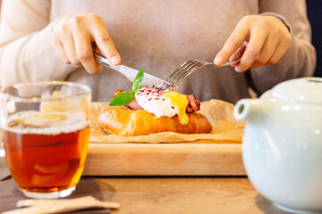 Woman has breakfast in a restaurant, egg benedict with croissant on a wooden plate under baking paper, brunch food style