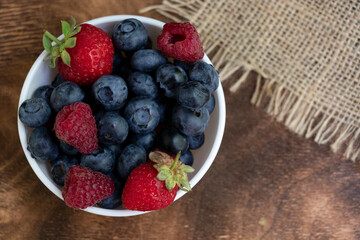 Set of summer berries in a small white bowl on a napkin and wooden background. Blueberries, raspberries, and strawberries are a varied healthy snack. Yummy.