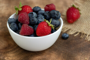 Set of summer berries in a small white bowl on a napkin and wooden background. Blueberries, raspberries, and strawberries are a varied healthy snack. Yummy.