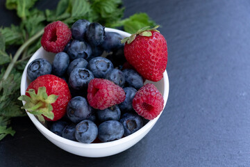 Set of summer berries in a small white bowl on a blackboard. Blueberries, raspberries, and strawberries are a varied healthy snack. Yummy.