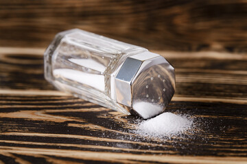 Some salt spilled out of the glass salt shaker with a silver metal lid lies on its side on a dark wooden table. Low key photography. - obrazy, fototapety, plakaty