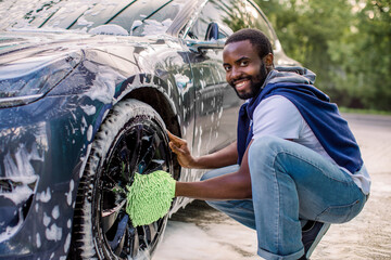 Side view of handsome young African man using green microfiber car wash mitt for cleaning rims of...