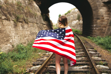 Young girl walking on the railway lines with the American flag on her back. 4th of July. Independence Day.
Travel, adventure, immigration and lifestyle concept.
