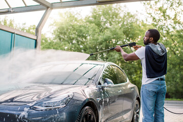 African stylish young focused man in casual clothes washing his modern electric car with a water...