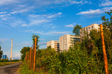 New residential buildings behind lush foliage and wire fence, bright blue sky and heating plant in background