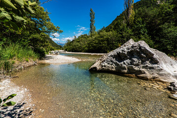Turquoise water of the french Drome river running between pebbly river banks and green forest with high mountains on the background.