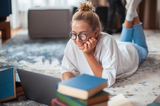Cute teen girl student in casual clothes, glasses lying on floor at laptop with books. Back to school. Exam preparation