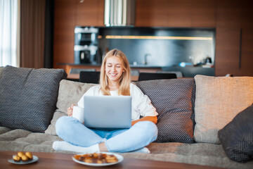 Smiling young girl sitting on sofa in Lotus position with laptop watching series with sweets and tea. Cozy home photo