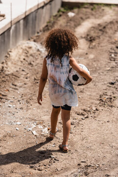 Back View Of Poor African American Child Holding Soccer Ball On Dirty Road Of Slum