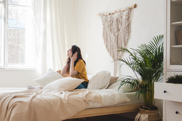 Obraz premium Young beautiful girl student in an orange T-shirt and blue jeans sits on bed with pillows and listens to an audiobook, music, podcast on headphones. Self-education concept at home during quarantine.