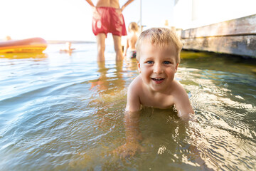 Fototapeta na wymiar Cute adorable cheerful caucasian little blond toddler boy enjoy having fun playing at lake or river beach water on warm sunset evening time outdoors.. Happy childhood vacation at countryside concept