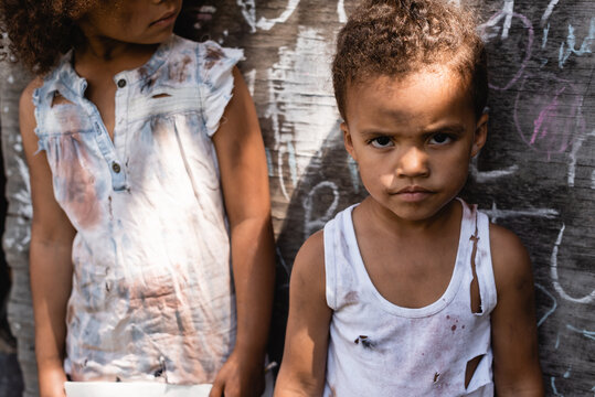 Poor African American Kid In Torn Clothes Standing With Brother Near Chalkboard