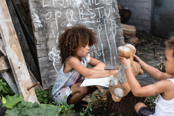 selective focus of curly african american kid writing in notebook near poor brother playing with soft toy