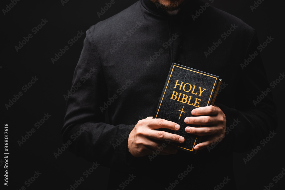 Wall mural cropped view of priest holding holy bible in hands isolated on black - Wall murals