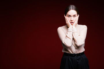 Surprised young woman on a red background presses her palms to her cheeks and looks at the camera. The student is surprised at the approach of the exam.