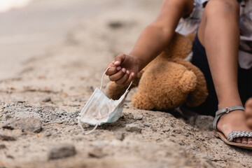 Fototapeta na wymiar cropped view of poor african american child touching dirty medical mask near teddy bear on ground