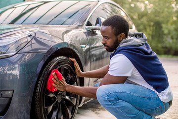 Obraz na płótnie Canvas Car washing on open air. Young hipster African bearded man cleaning a wheel, car rims of modern luxury gray electric car with red microfiber cloth, at outdoor car wash self service.